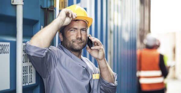 A construction worker with a hard hat is leaning against a wall and talking on the phone. A worker in a reflective vest is in the background.