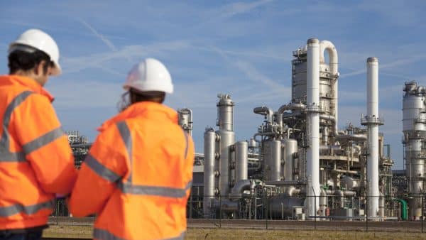 two oil and gas plant employees in hard hats conversing with each other. In the background is a large oil and gas plant.