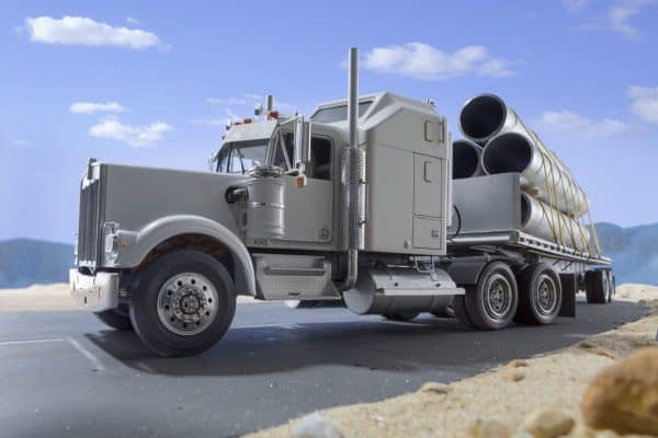 A silver semi-truck loaded with large industrial pipes secured by yellow straps drives on a highway.