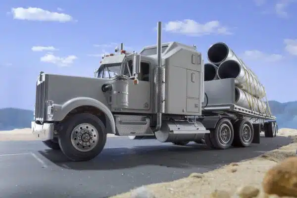 A silver semi-truck loaded with large industrial pipes secured by yellow straps drives on a highway.