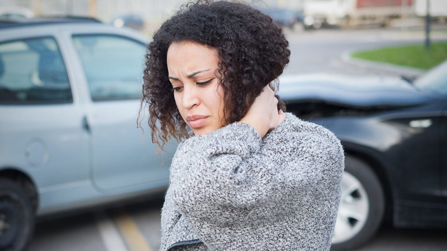 A woman is holding her neck as if in pain. A wrecked vehicle can be seen in the background.