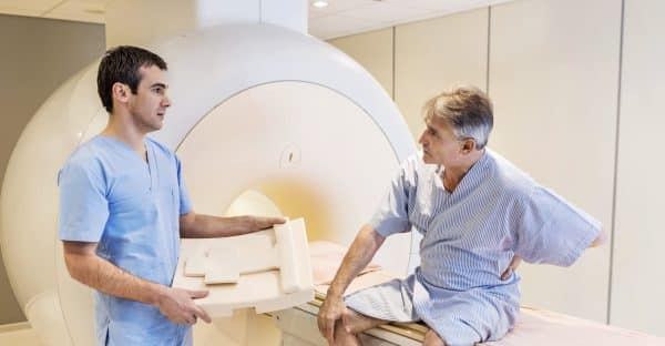A man in hospital robes holding his back sits on an MRI machine and is speaking to his doctor.