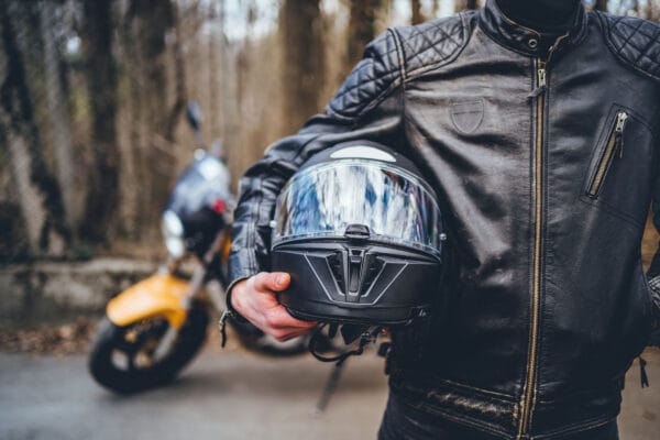 A motorcycle rider in a leather jacket carries a full-face helmet. Their motorcycle can be seen in the background.