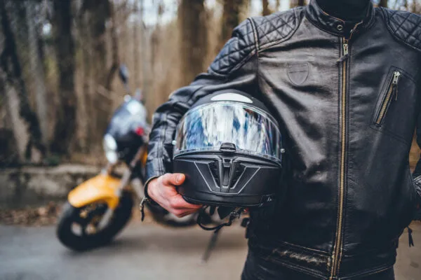 A motorcycle rider in a leather jacket carries a full-face helmet. Their motorcycle can be seen in the background.