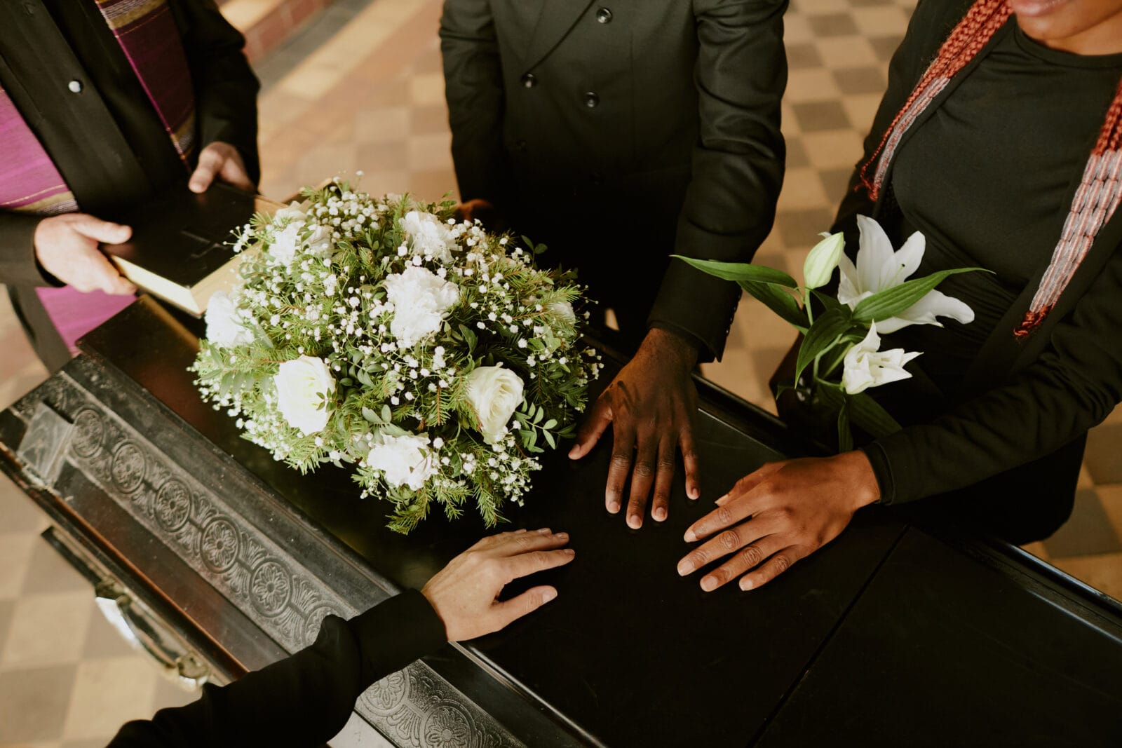 Hands on a black casket topped with flowers. A funeral officiant holds a Bible.