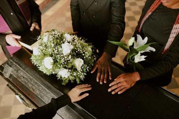 Hands on a black casket topped with flowers. A funeral officiant holds a Bible.