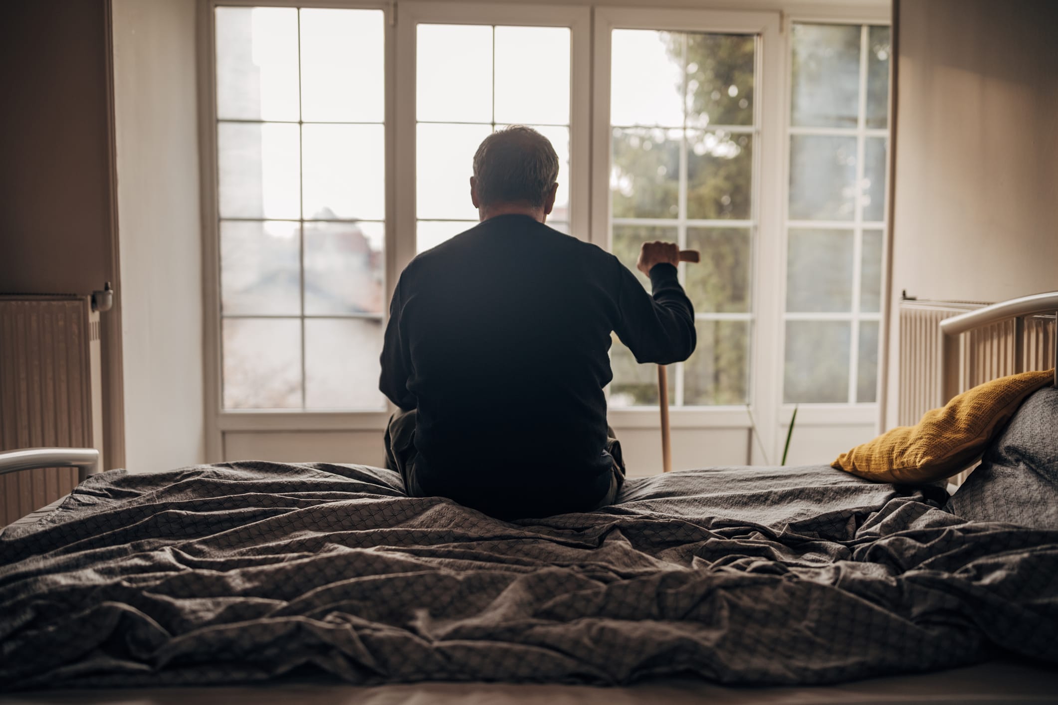 An elderly man with a cane, sitting on a bed and looking out the window.