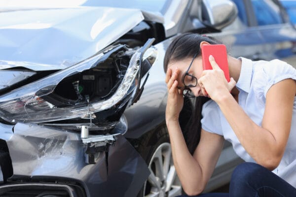 A distressed woman talks on the phone while sitting next to her damaged car.