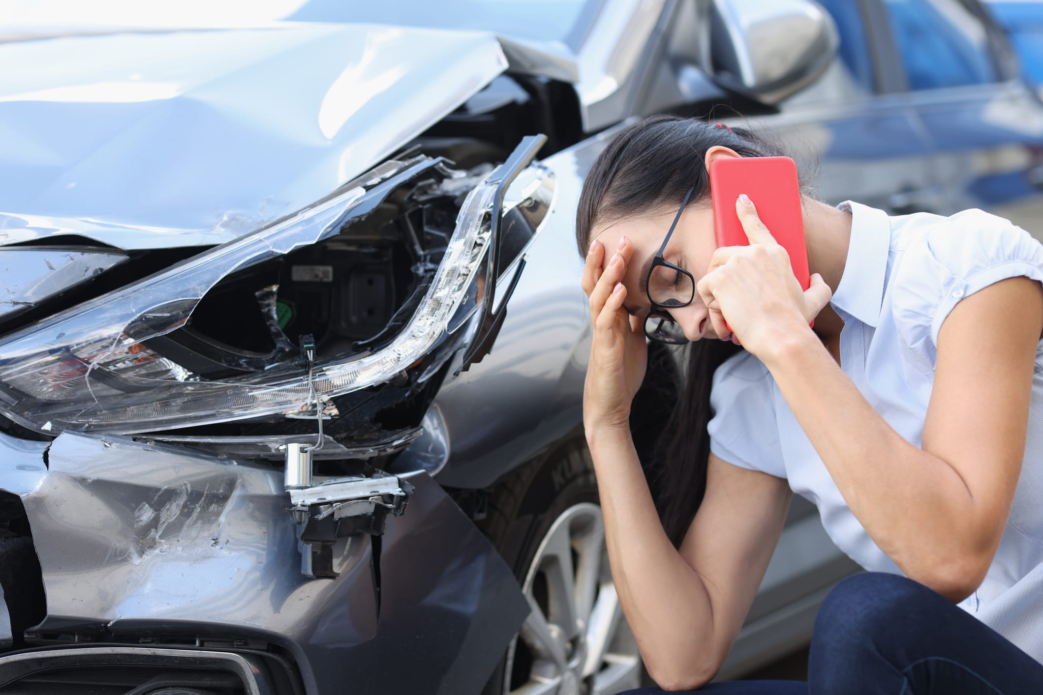 A distressed woman talks on the phone while sitting next to her damaged car.