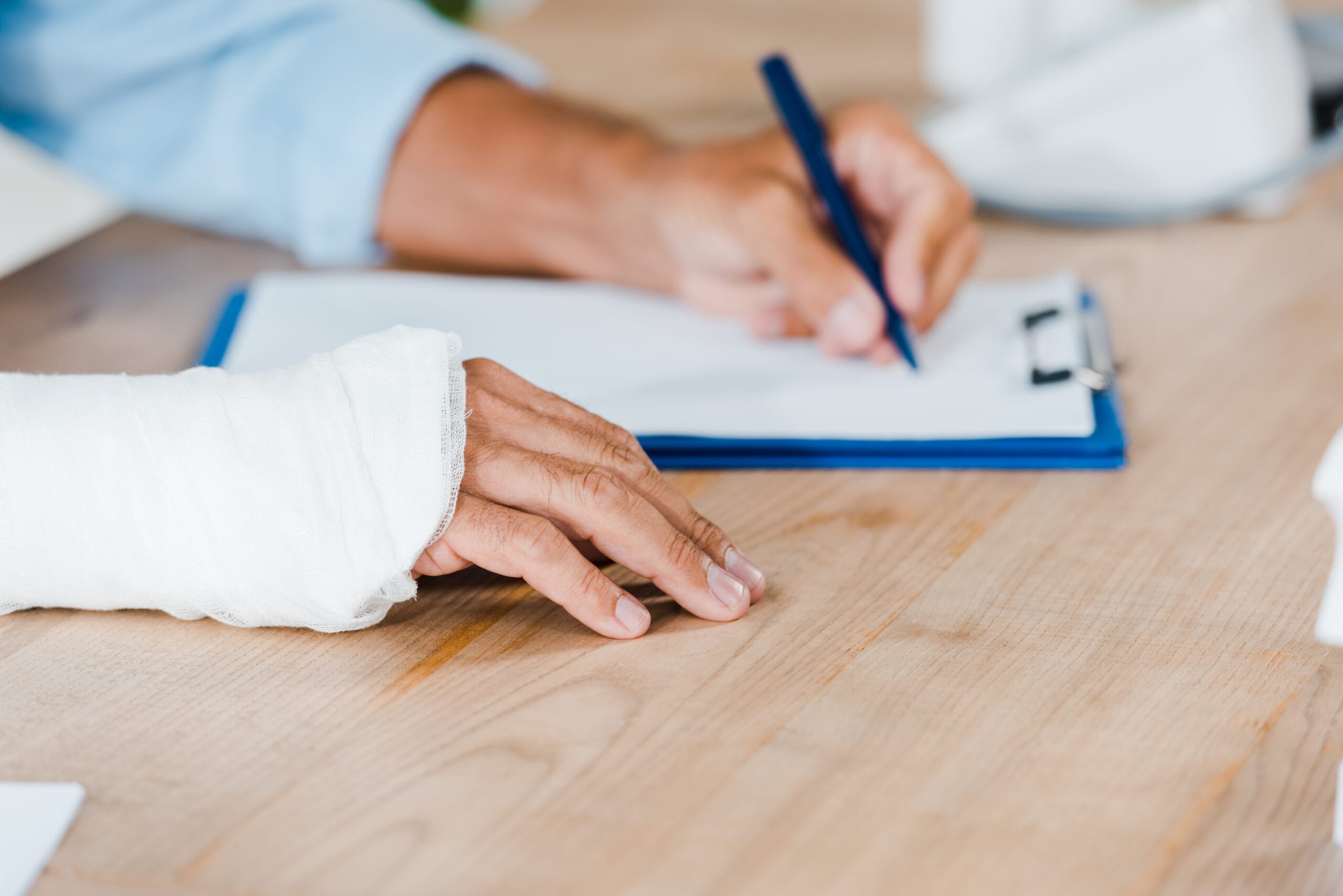 Person with a bandaged hand resting on a table while signing paperwork on a clipboard with a pen.