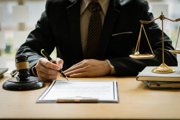 A workers’ comp lawyer fills out a document with a pen. Scales, a stack of books, and a gavel and block all sit on the desk in front of him.