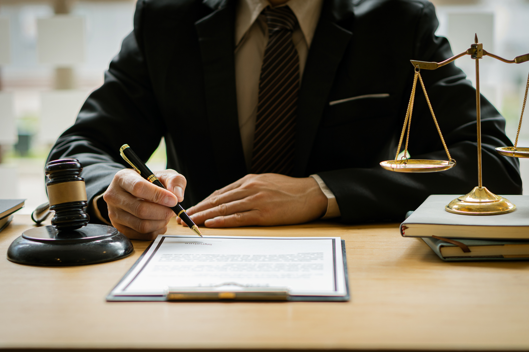 A workers’ comp lawyer fills out a document with a pen. Scales, a stack of books, and a gavel and block all sit on the desk in front of him.