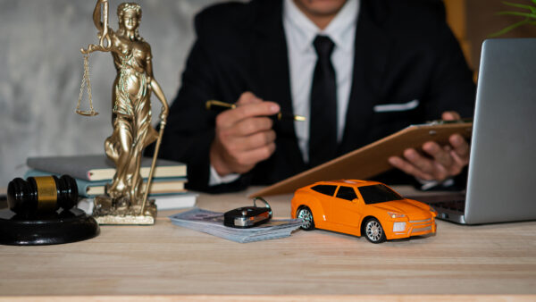 A car accident lawyer sits at his desk holding a clipboard and pen. A computer, a stack of books, a Lady Justice statuette, a gavel and block, a stack of money, and an electronic key fob are sitting on the desk in front of him.