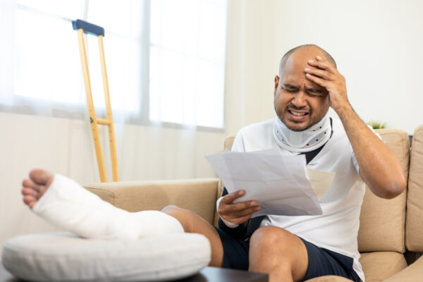 A man with his leg in a cast reads his medical bills while holding his head in distress. He sits on a couch with his crutch leaned against the window next to him.