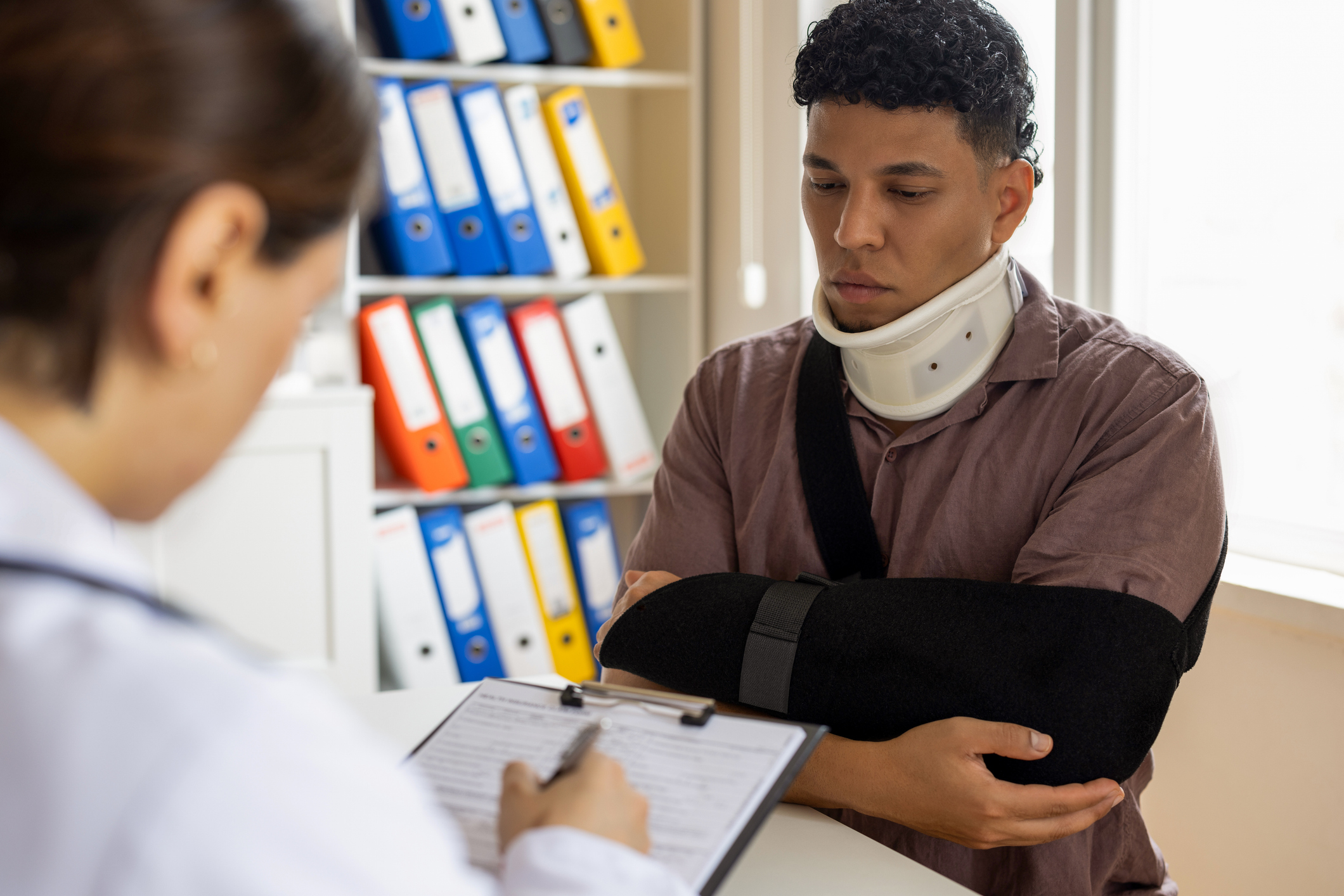An injured worker with a neck brace and their arm in a sling sits in front of a medical professional who is taking notes on a clipboard. Different-colored binders fill the shelf next to them.
